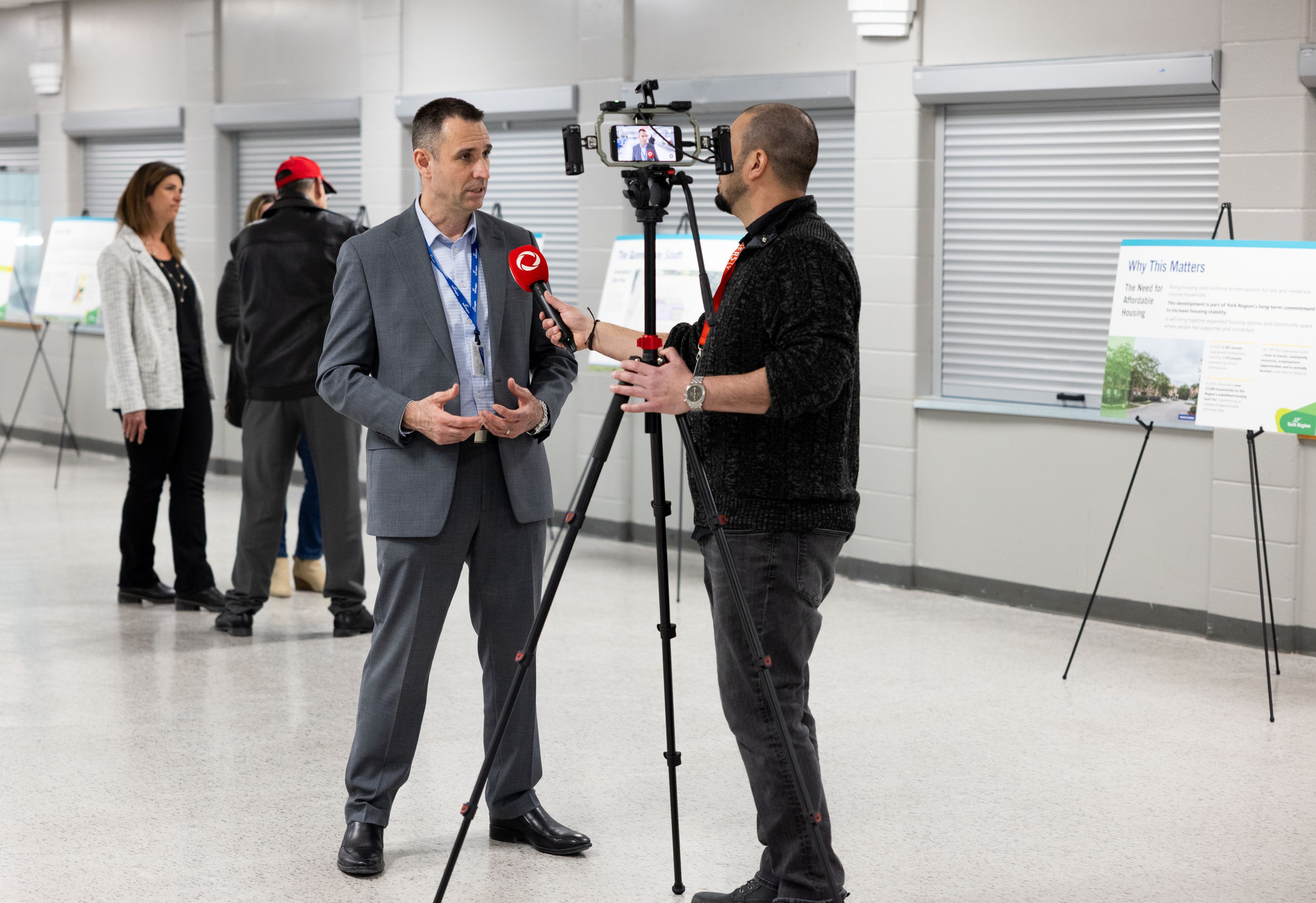 A person is being interviewed on camera at The Queensway South community open house, with display boards and attendees in the background.