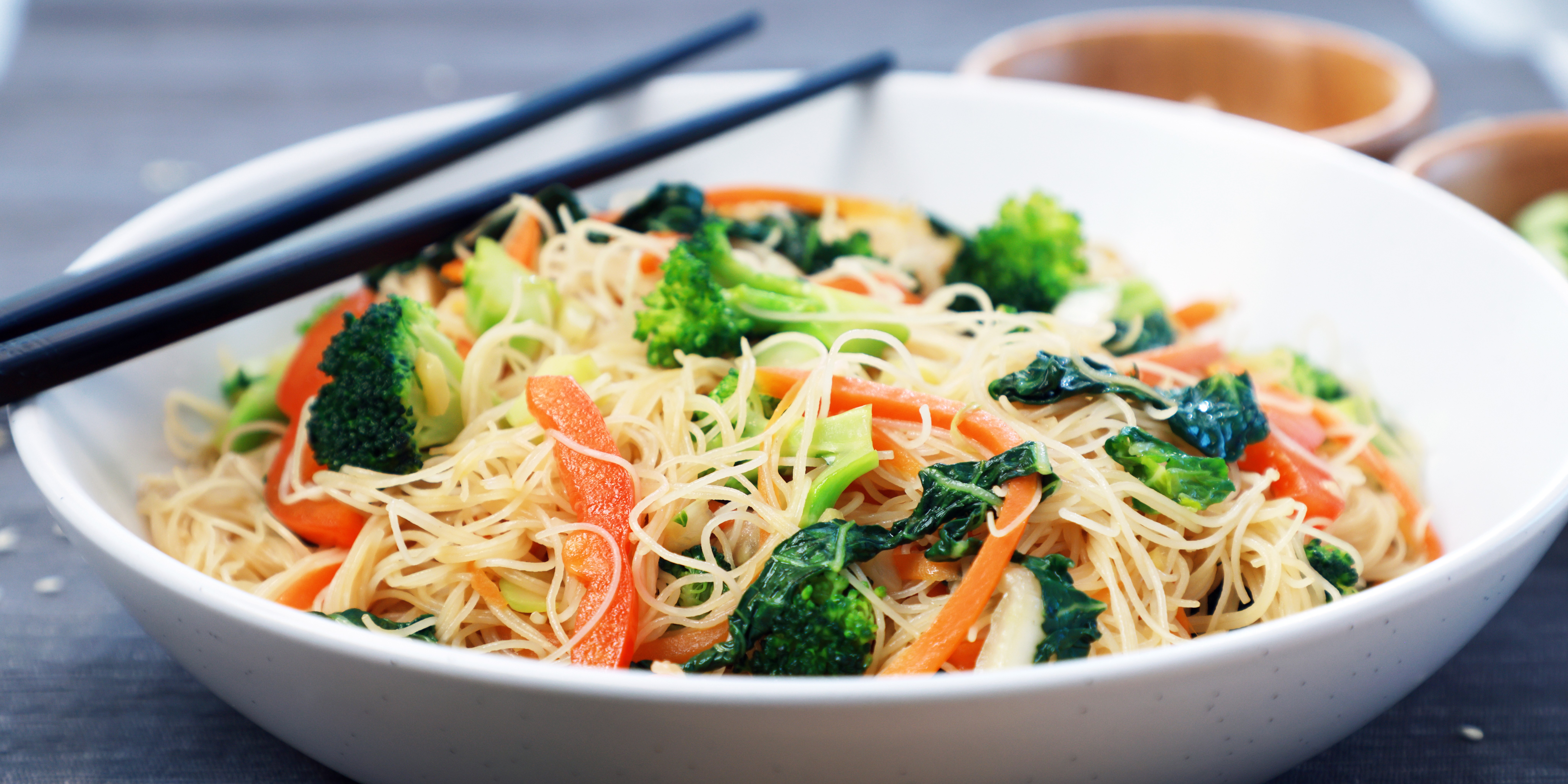 Rice vermicelli noodles stir‑fried with broccoli, carrots, and greens in a white bowl with chopsticks.