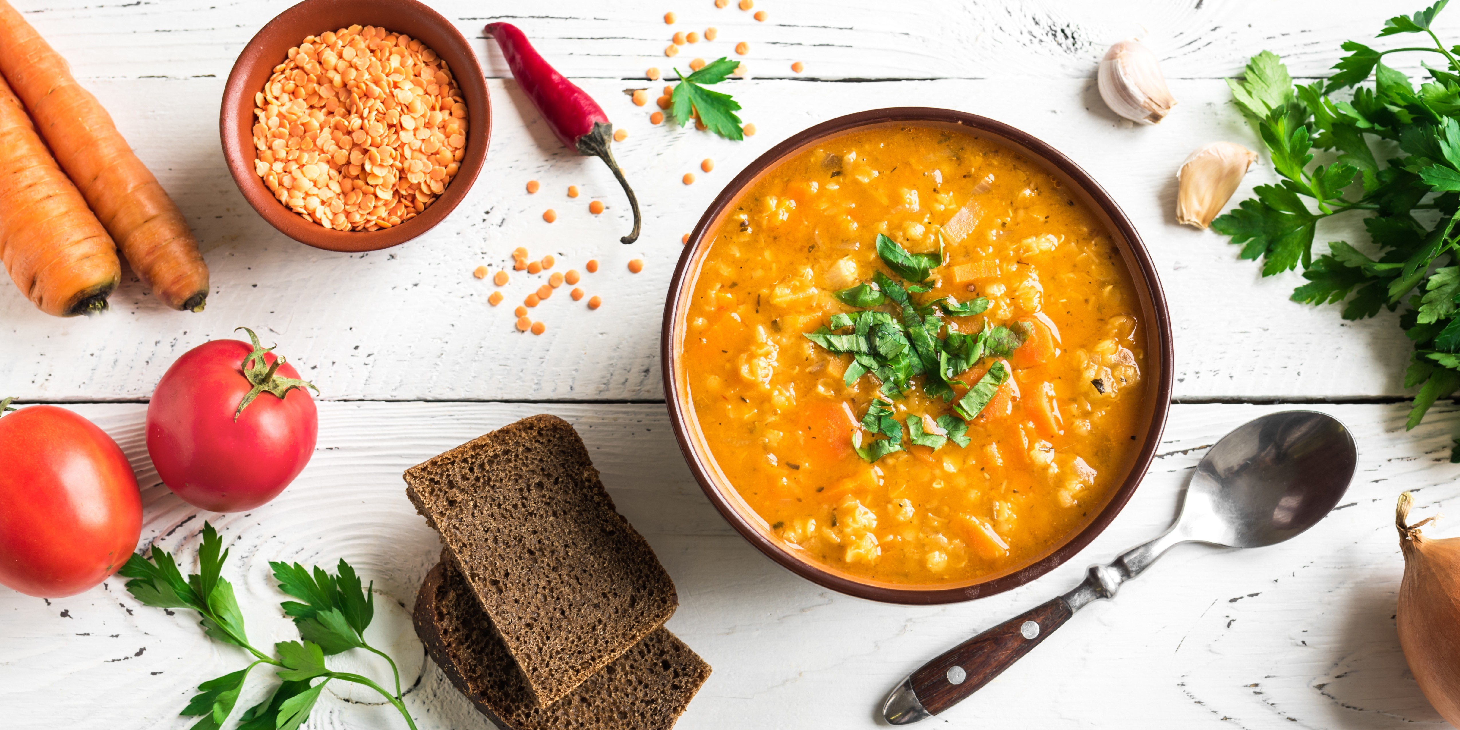 Bowl of red lentil soup with herbs, surrounded by fresh ingredients and bread.