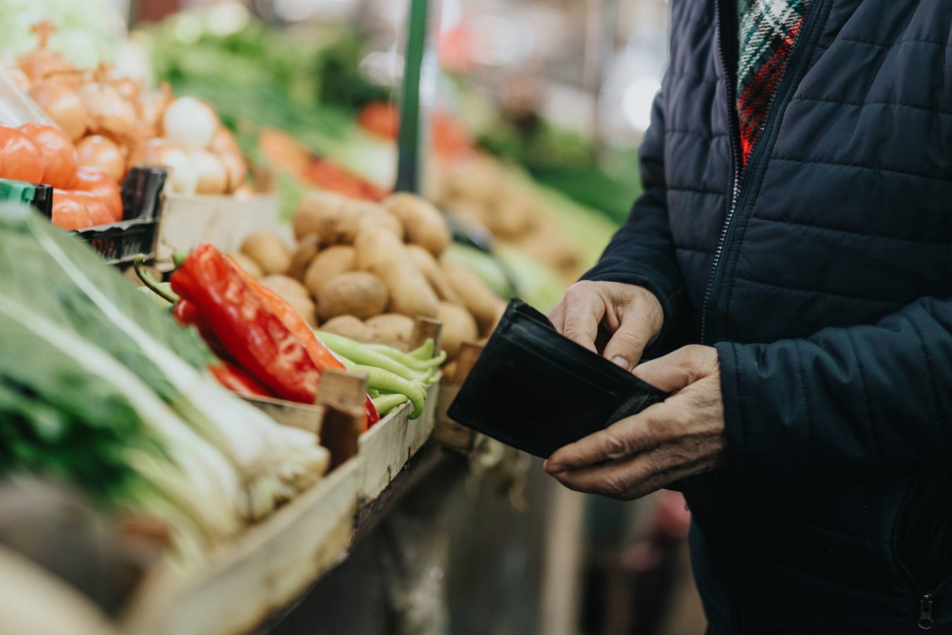 Person holding a wallet while standing beside a display of fresh vegetables at a grocery store.