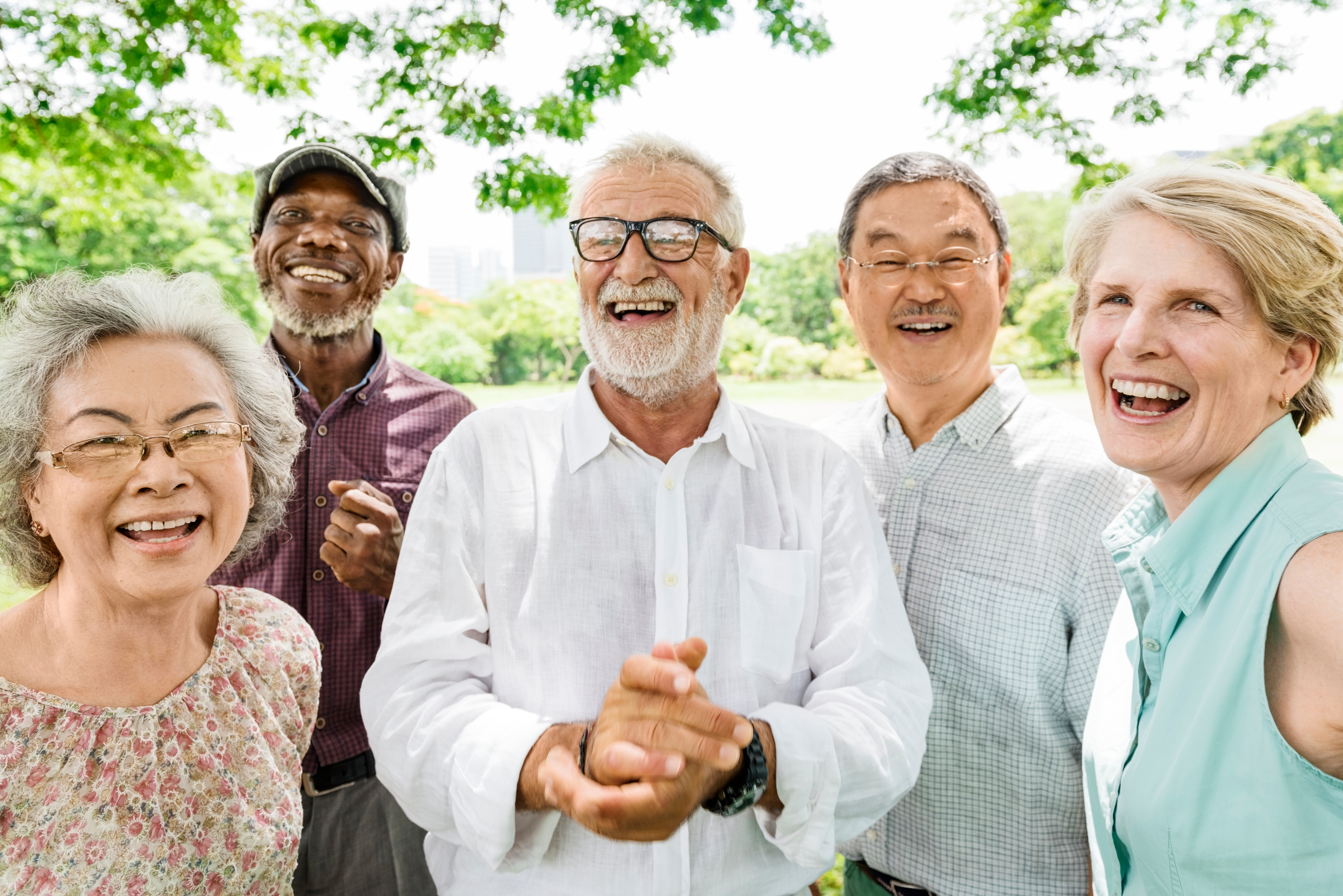 A diverse group of five older smiling adults gathered outdoors in a park, standing close together and facing the camera with trees in the background.