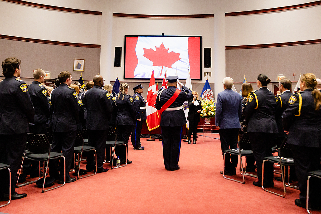 Uniformed paramedics stand in rows facing the Paramedic Graduation ceremony, with one person saluting and Canadian flags displayed in front of a large screen showing a red maple leaf flag.