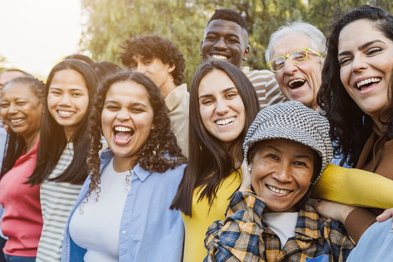 Multicultural and multi-generational group of nine smiling people; trees and sunshine are in the background