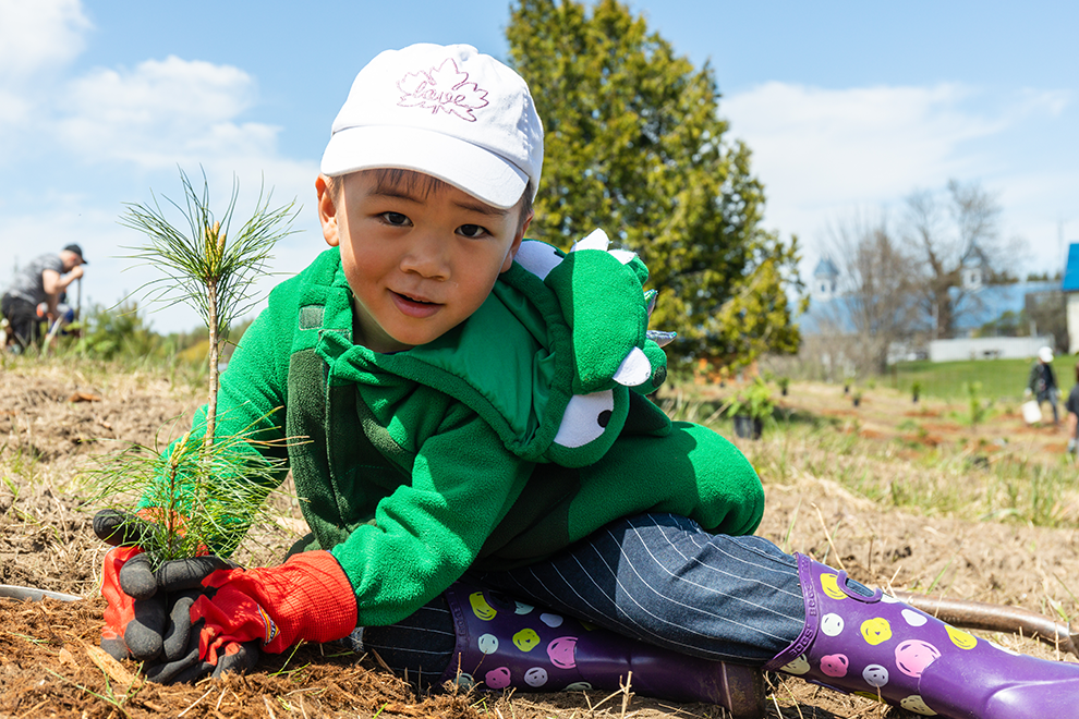 a young child dressed in a hat and green jacket planting a pine seedling