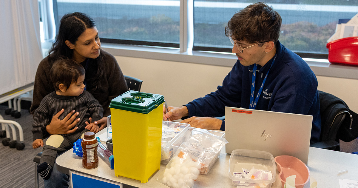 Healthcare worker at a table with medical supplies speaks to a caregiver holding a child in a clinic setting.