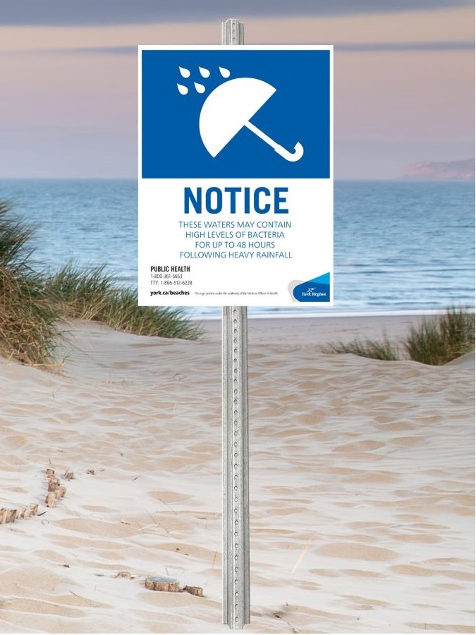 York Region Beach Water Notice Sign with an umbrella icon on a blue background. On a white background, the sign states: &quot;Notice. These waters may contain high levels of bacteria for up to 48 hours following heavy rainfall. Public Health: 1-800- 361-5653; TTY: 1-866-512-6228; york.ca/beaches&quot;